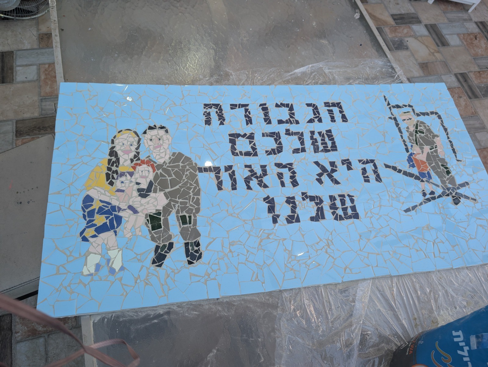 Handmade mosaic mural of a family and a soldier standing in a doorway, with Hebrew lettering