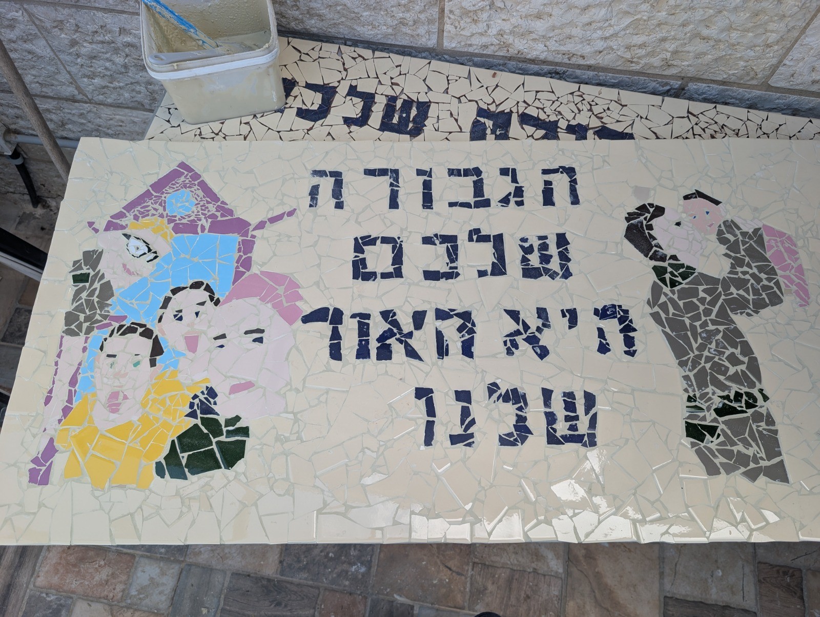 Handmade mosaic mural of family faces and a soldier lifting a child, with Hebrew lettering