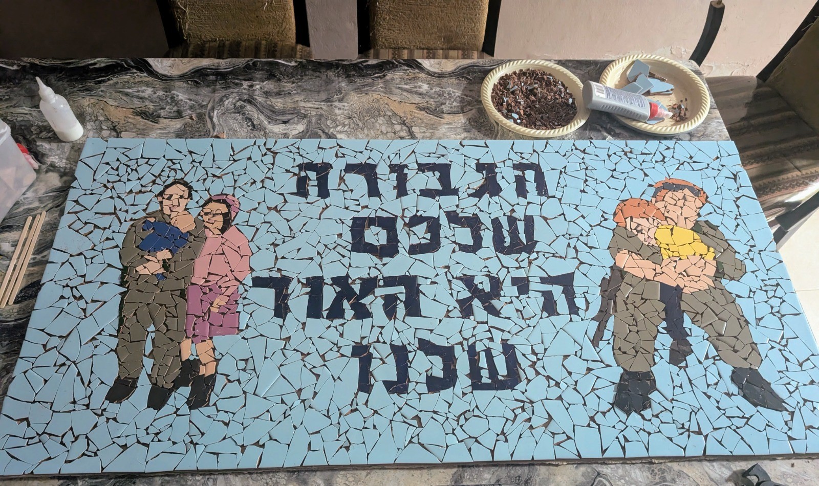 Handmade mosaic mural of a soldier embracing a woman and a soldier holding a child, with Hebrew lettering
