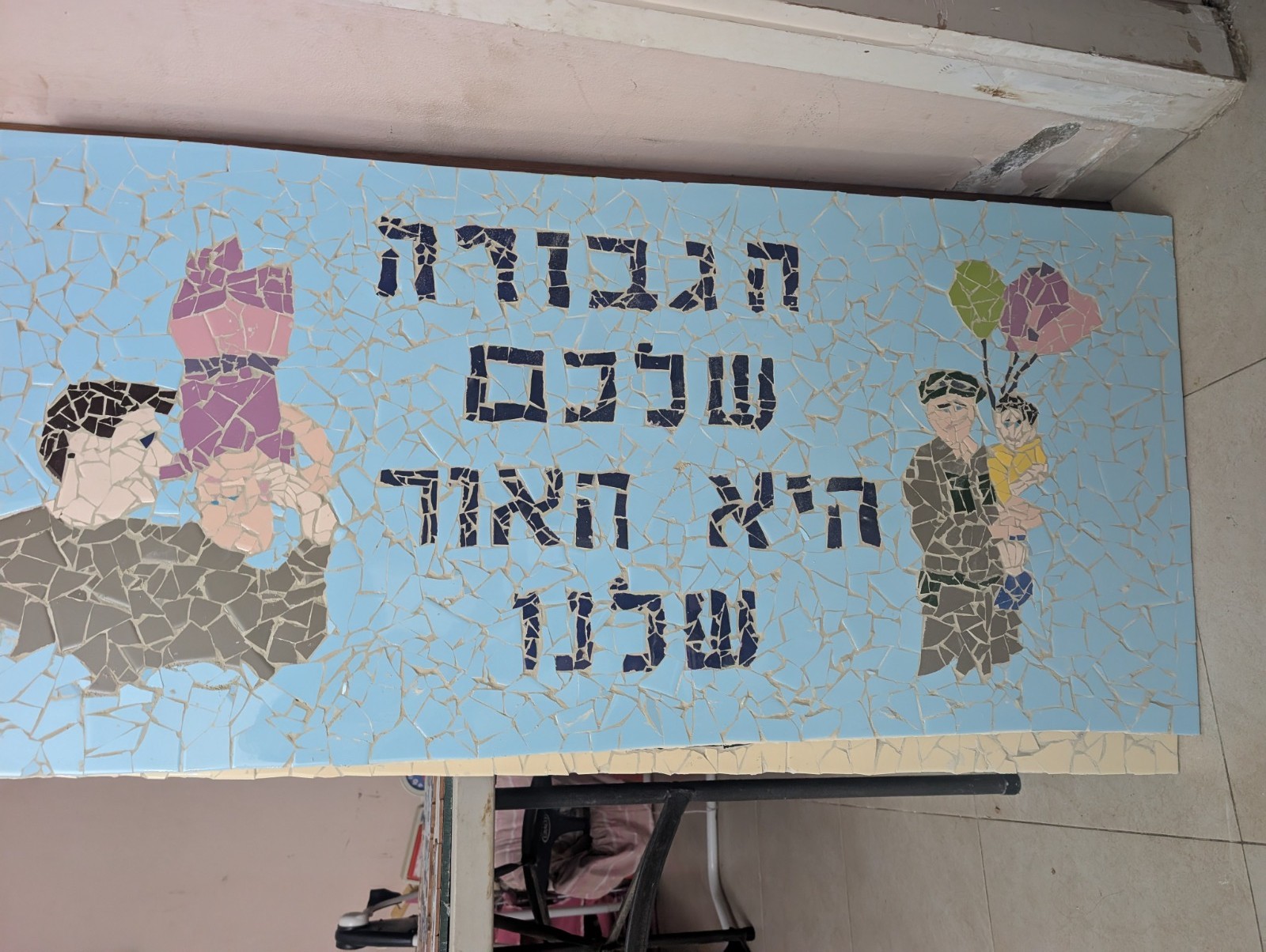 Handmade mosaic mural of a soldier lifting a child and a child with balloons, with Hebrew lettering