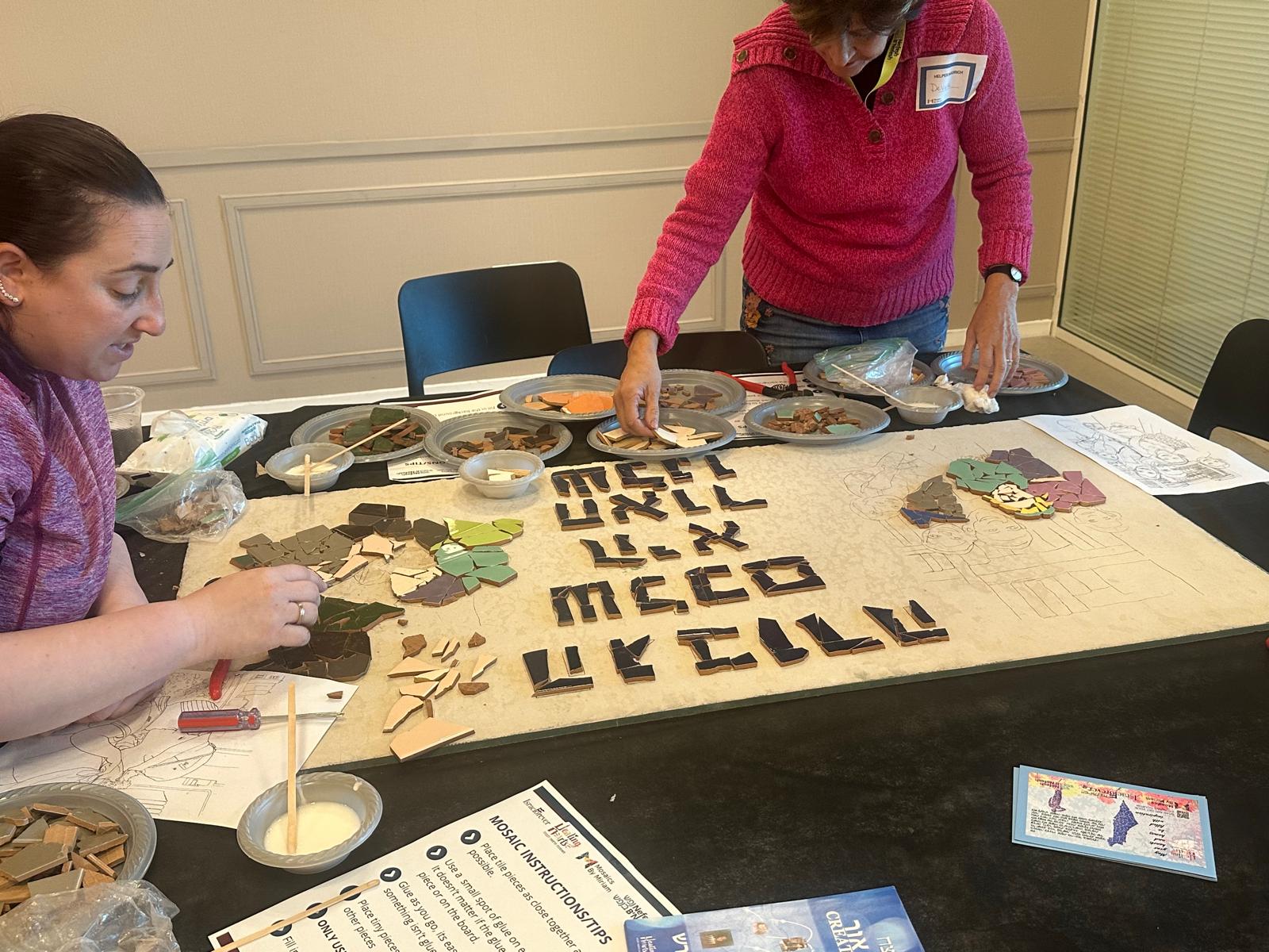 Two women placing tiles on a large mural design at a workshop table