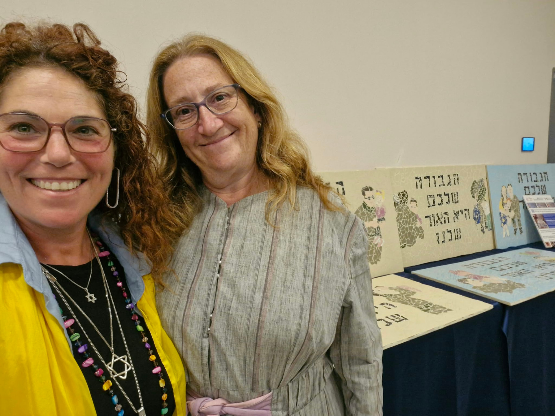 Dr. Elana Heideman and Miriam Gottlieb beside mosaic tribute murals at an exhibition