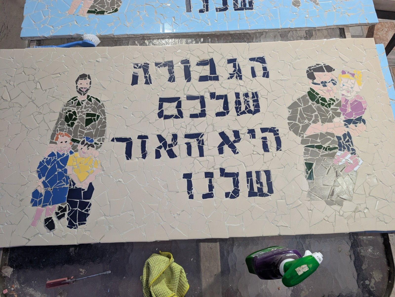 Handmade mosaic mural of a soldier with children and Hebrew lettering, lying on a workshop table